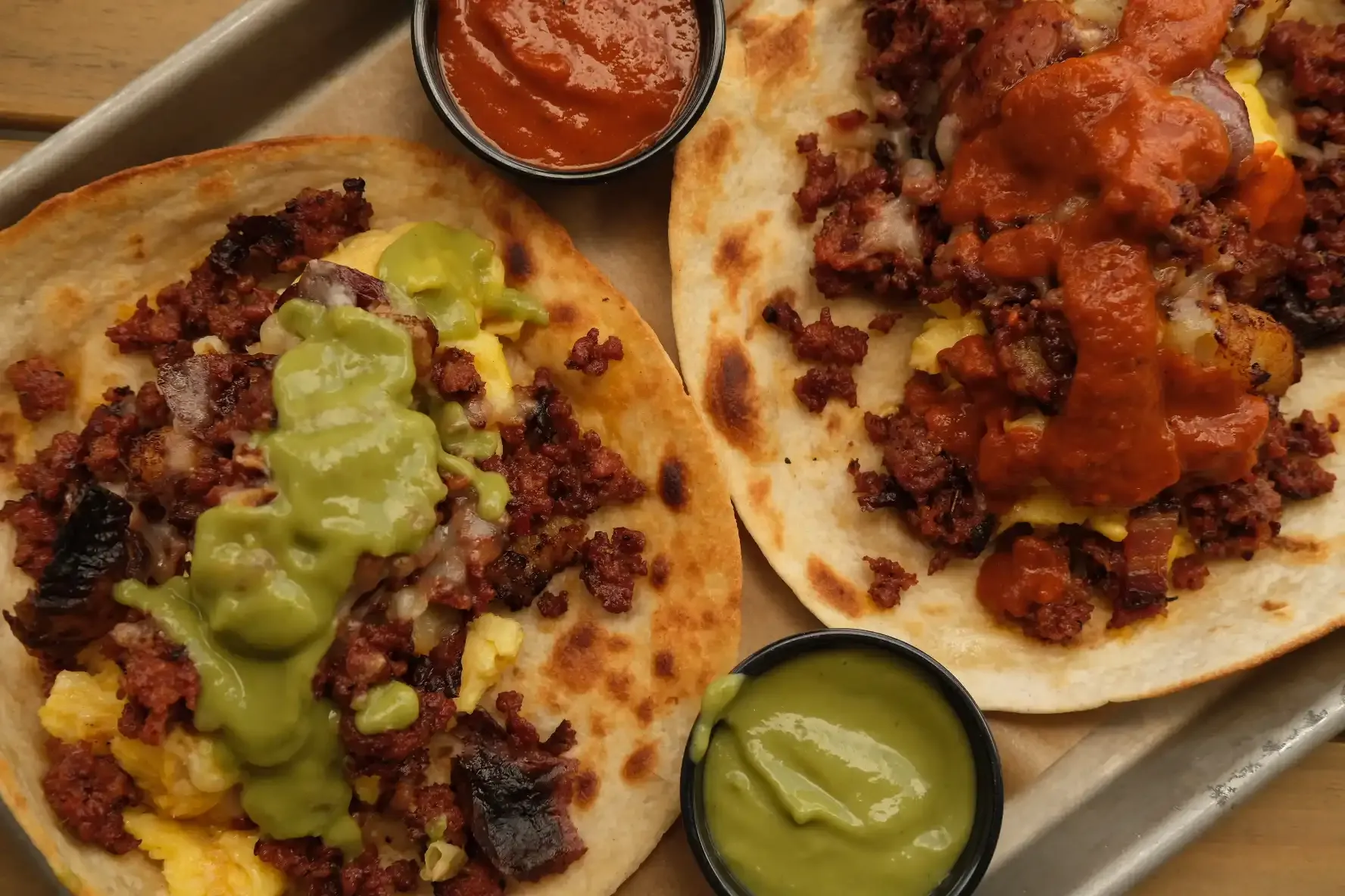 Chorizo and egg tacos on griddled flour tortillas, one topped with salsa verde and one with salsa roja, with a cup of each salsa