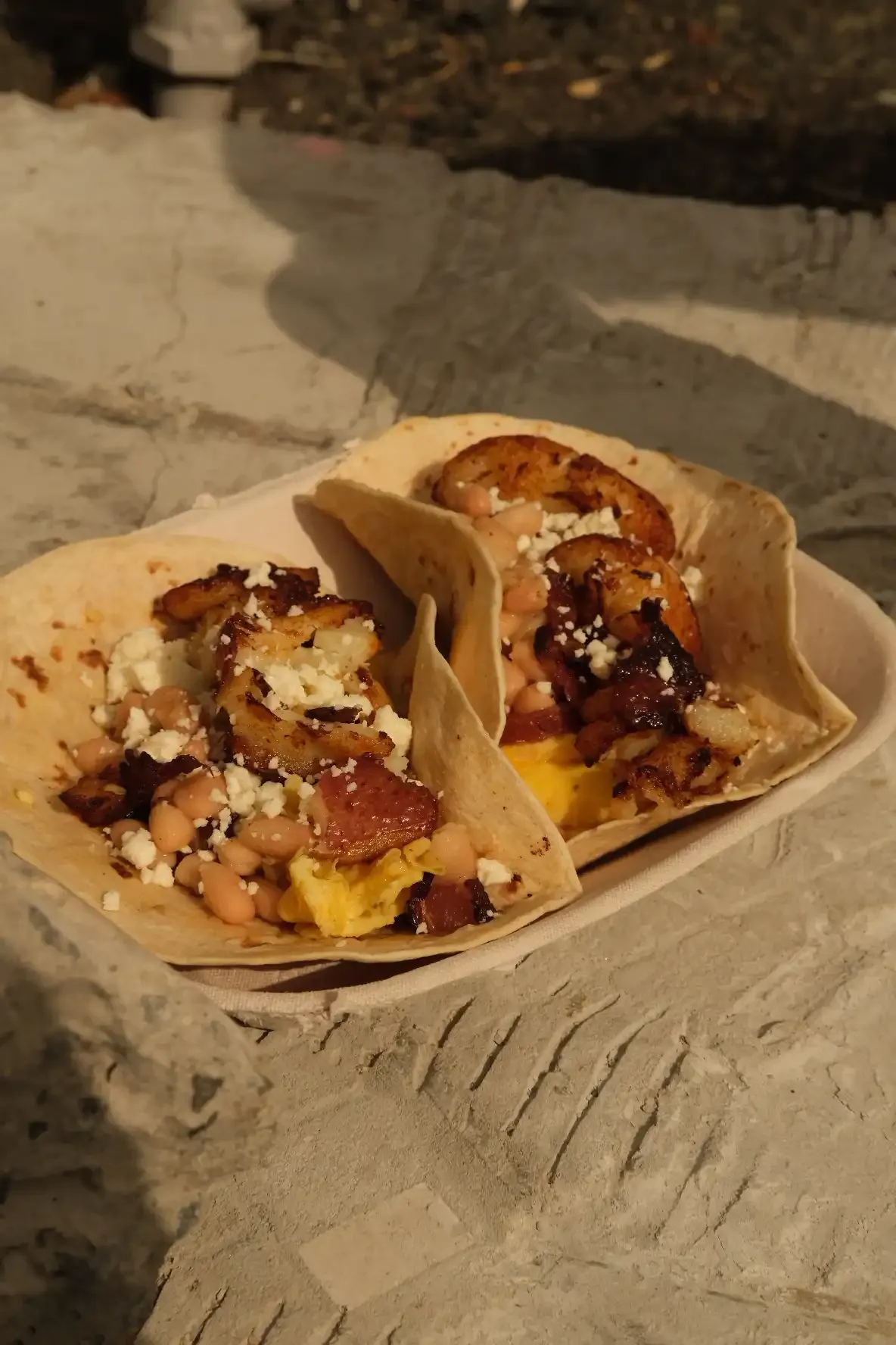 Two breakfast tacos in a paper boat on a stone surface, filled with scrambled egg, white beans, bacon, crispy potato and queso fresco