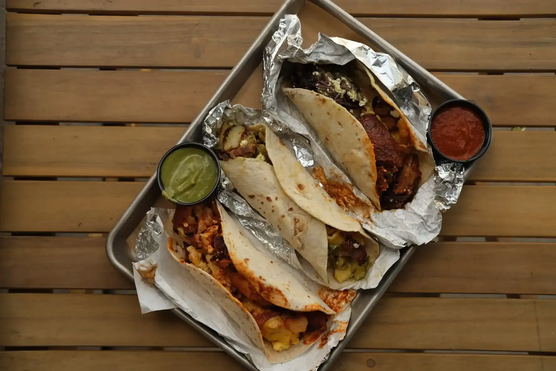 Three foil-wrapped breakfast tacos on a metal tray with cups of salsa verde and salsa roja