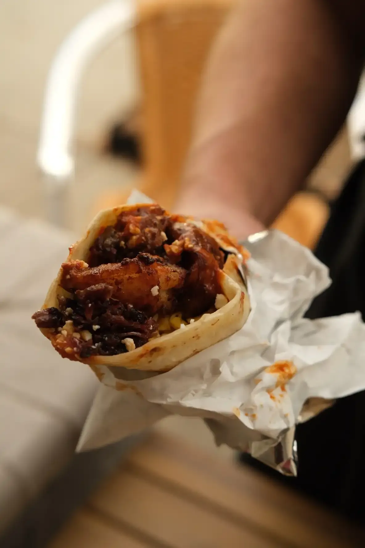 A hand holding a foil-wrapped breakfast taco filled with red-sauced meat and potato, scrambled egg and queso fresco